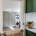 View from a dark green kitchen through a doorway into a sage utility room with a stacked washing machine and tumble dryer