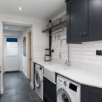 Galley utility room with navy shaker cabinetry, a Belfast sink, white metro tiles and two washing machines beneath a quartz worktop