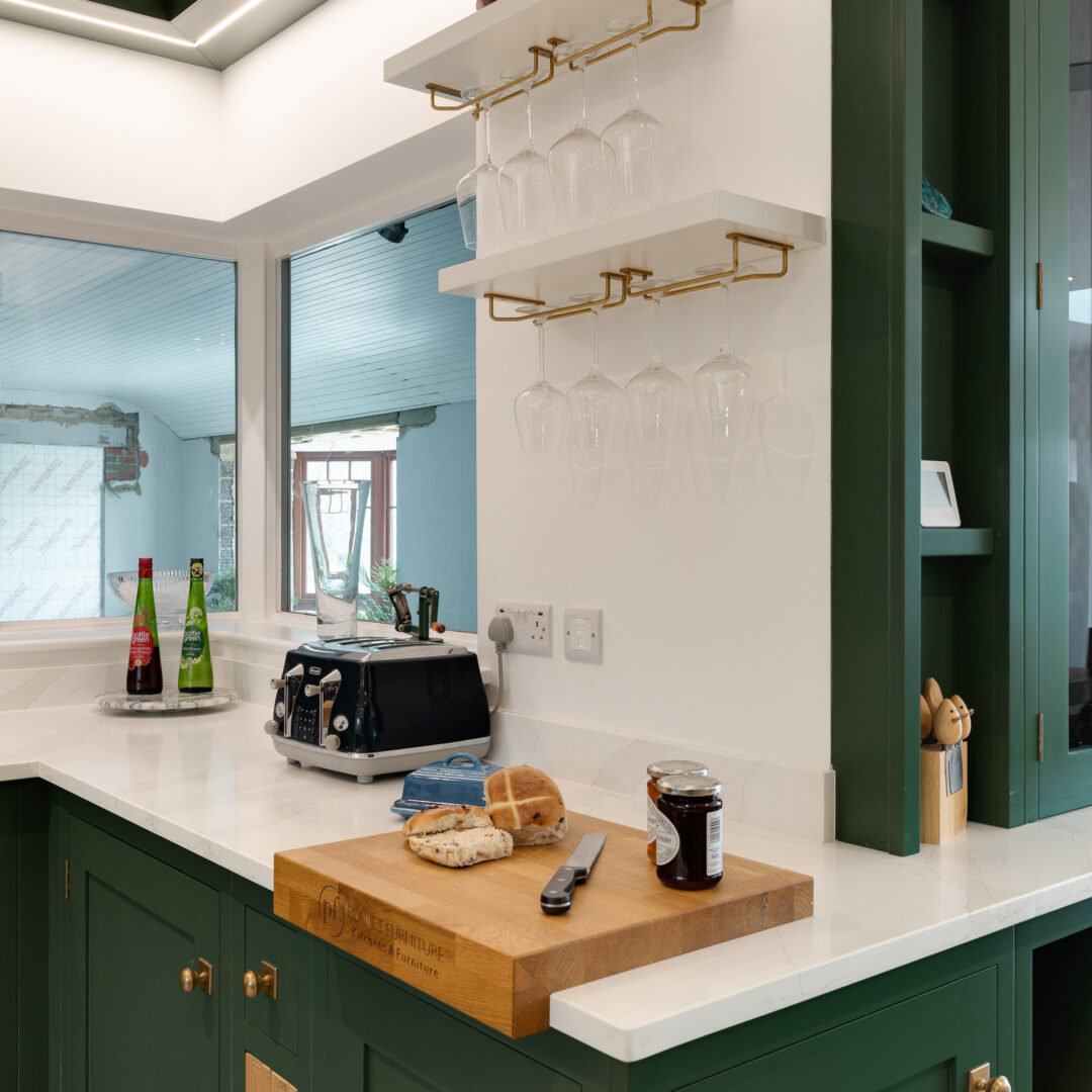 Brass hanging wine glass racks beneath white floating shelves above a dark green in-frame shaker kitchen