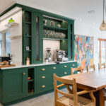 Dark green in-frame shaker kitchen with a dresser, plate rack and coffee station, open to a dining area with a long oak table