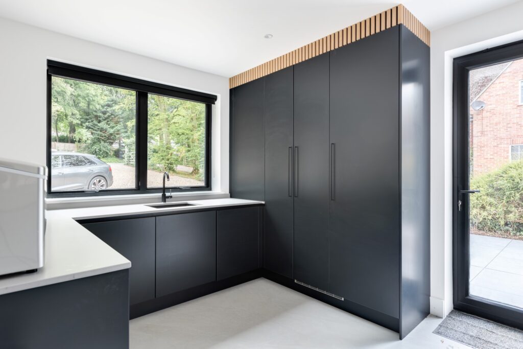 Charcoal handleless utility room with full-height storage, a slatted oak detail above and an L-shaped worktop beneath the window