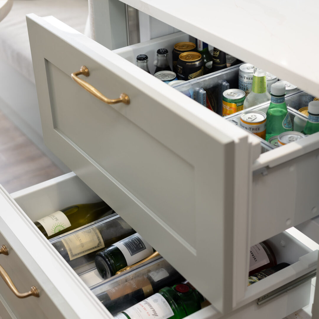 Two refrigerated drawers open in a pale grey shaker island with antique brass handles, holding jars, tins and wine bottles